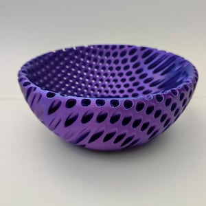 Puede incluir: Un cuenco decorativo morado con un diseño perforado único. El cuenco tiene forma redondeada con una superficie texturizada y un interior morado oscuro. El exterior presenta una serie de cortes ovalados. Diámetro aproximado de 15 cm.