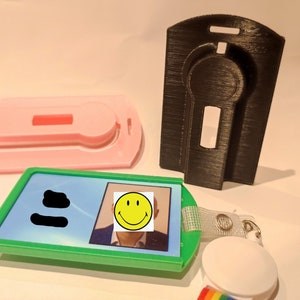 Può includere: Un porta badge verde con foto e faccina sorridente, un porta badge rosa e un porta badge nero. Il porta badge ha un cordino bianco e una clip arcobaleno.