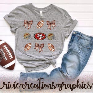 May include: A gray t-shirt with football-themed graphics, including footballs, bows, and helmets, alongside a San Francisco 49ers logo. A brown and white football and blue jeans complete the arrangement.