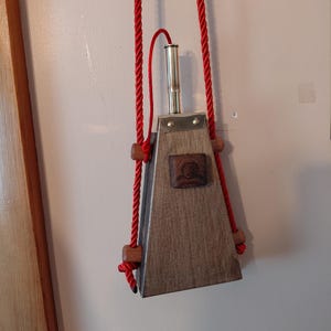 Puede incluir: Un frasco de pólvora de madera con forma trapezoidal y una boquilla metálica en la parte superior. El frasco presenta un emblema cuadrado de madera oscura en el frente, asegurado por dos pequeños remaches metálicos. Cuelga de una cuerda roja.