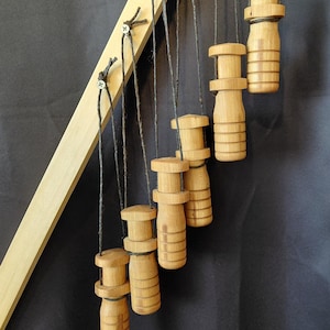 May include: A series of seven wooden objects, each suspended by a black cord from a light-colored wooden frame. The objects are cylindrical with a rounded top and bottom, and a series of horizontal grooves. The background is a dark gray.