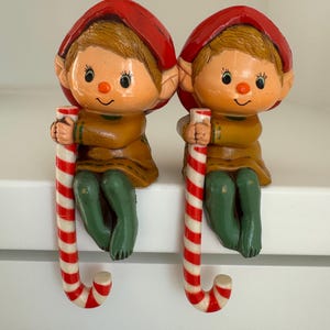 May include: Two ceramic elf figurines with red hats, holding candy canes. The elves have tan skin, brown hair, and are dressed in brown and green outfits. The candy canes are red and white striped.