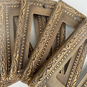 May include: A collection of ornate, antique-style light switch covers. The covers are made of a bronze-colored metal with detailed, raised floral and geometric patterns. Each cover has a single rectangular opening and screw holes.