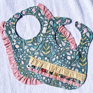 May include: Two baby bibs featuring a dark teal fabric with a botanical print of leaves, berries, and flowers. The bibs are accented with pink and yellow ruffled trim along the edges and include pink plastic snap closures at the neck.