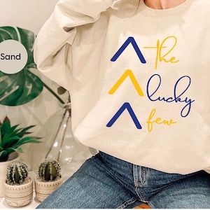 Op de afbeelding: Een crèmekleurig sweatshirt met een grafisch ontwerp van drie blauwe en gele pijlen die omhoog wijzen. De tekst "the lucky few" is geschreven in een cursief schrift onder de pijlen.