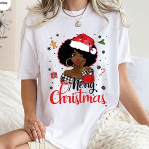 Könnte beinhalten: Weißes T-Shirt mit einem festlichen Aufdruck einer Frau mit Weihnachtsmütze. Das Design enthält die Worte "Merry Christmas" in Rot, sowie Schneeflocken, einen Lebkuchenmann und eine Zuckerstange.