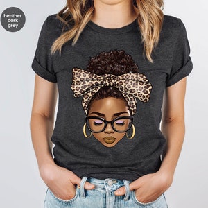 May include: A heather dark grey t-shirt with a graphic of a woman with brown hair in a bun wearing a leopard print headband and glasses.