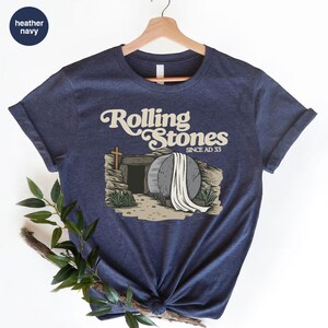 May include: A heather navy blue t-shirt with a graphic of a stone tomb with a white sheet draped over it. The text "Rolling Stones Since AD 33" is above the tomb.
