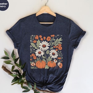 Puede incluir: Una camiseta azul marino jaspeado con un estampado gráfico central de margaritas blancas, caléndulas naranjas y dos calabazas naranjas rodeadas de follaje verde. La camiseta tiene mangas cortas y un cuello redondo.