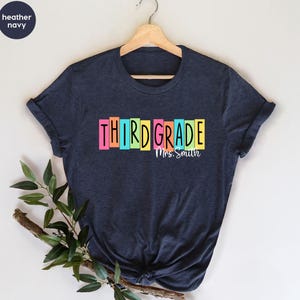 May include: A heather navy short-sleeve t-shirt featuring the text "THIRD GRADE" in black letters, each set against a colorful rectangular block background. Below the text, the name "Mrs. Smith" is printed in a white cursive script font.