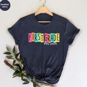 以下が含まれることがあります： ヘザーネイビーの半袖Tシャツ。FIRST GRADEの文字が黒でプリントされ、それぞれの文字がピンク、オレンジ、ティール、イエロー、ライトグリーンの縦長長方形のブロックの上に配置されています。ブロックの下にはMrs. Smithという白い文字がプリントされています。