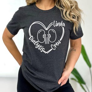 Könnte beinhalten: Ein dunkelgraues T-Shirt mit einem weißen Herzdesign. Das Herz zeigt zwei Nieren und den Text "Linda Dialysis Crew".