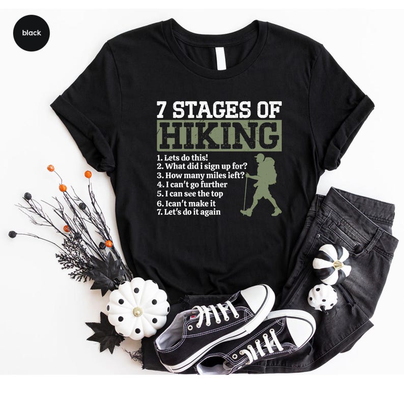 May include: A black short-sleeved t-shirt featuring the white text 7 STAGES OF HIKING and a list of seven numbered phrases describing the hiking experience, accompanied by a silhouette of a hiker with a backpack and a walking stick.