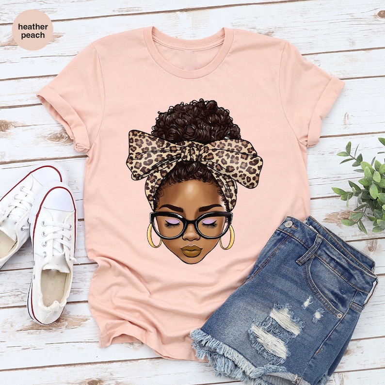 May include: A pink t-shirt with a graphic of a woman with brown skin, brown hair, and a leopard print headband. The woman is wearing glasses and has her eyes closed.