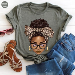 May include: A green t-shirt with a graphic of a woman with brown skin, brown hair, and closed eyes. She is wearing a leopard print headband and glasses.