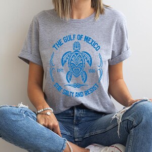Puede incluir: Una camiseta gris con un gr&aacute;fico azul de una tortuga marina y el texto "The Gulf of Mexico Est. 1550 Stay Salty and Resist".