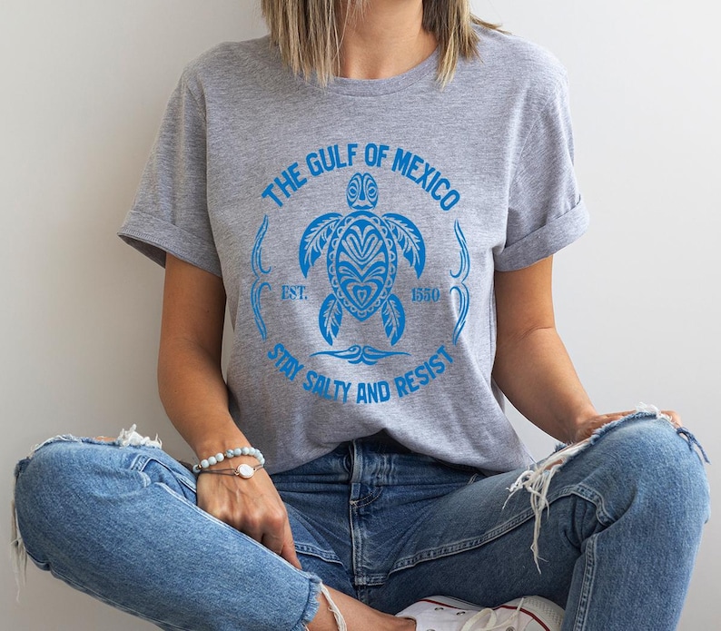 Puede incluir: Una camiseta gris con un gr&aacute;fico azul de una tortuga marina y el texto "The Gulf of Mexico Est. 1550 Stay Salty and Resist".