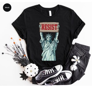 May include: Black t-shirt with a graphic of the Statue of Liberty holding a sign with the word "RESIST" in red. The shirt is styled with black jeans and black trainers. Decorative pumpkins and branches are also present.