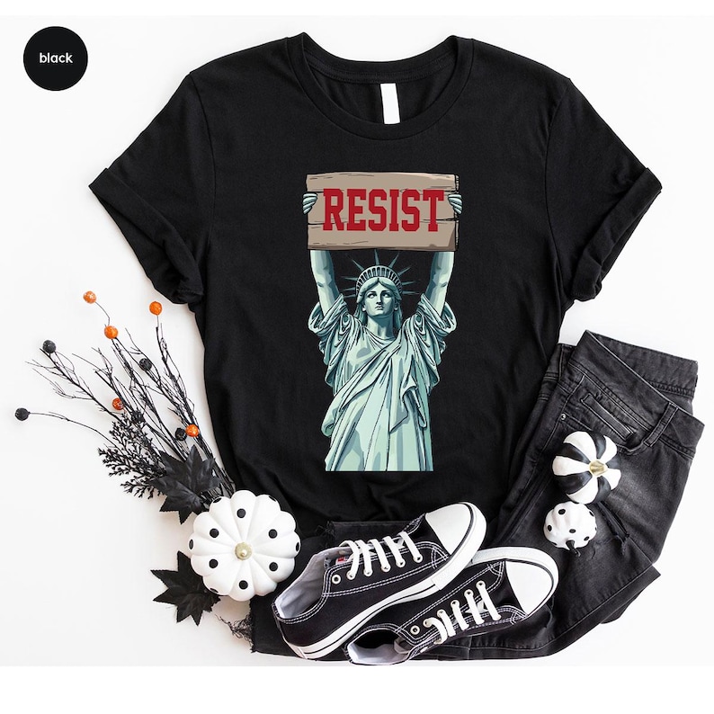 May include: Black t-shirt with a graphic of the Statue of Liberty holding a sign with the word "RESIST" in red. The shirt is styled with black jeans and black trainers. Decorative pumpkins and branches are also present.
