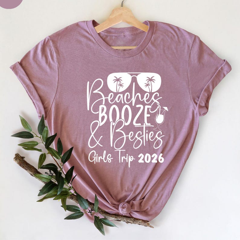 May include: Mauve t-shirt with the text "Beaches, Booze & Besties, Girls Trip 2026" in white script. The design includes sunglasses with palm trees and a cocktail glass. The shirt is knotted at the bottom and has rolled-up sleeves.