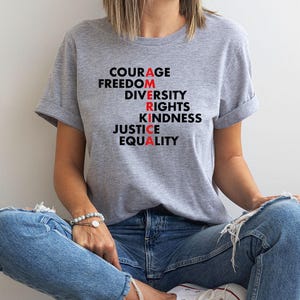 May include: A light gray t-shirt with the words "COURAGE, FREEDOM, DIVERSITY, RIGHTS, KINDNESS, JUSTICE, EQUALITY" printed in black and red. The shirt is worn with blue jeans.