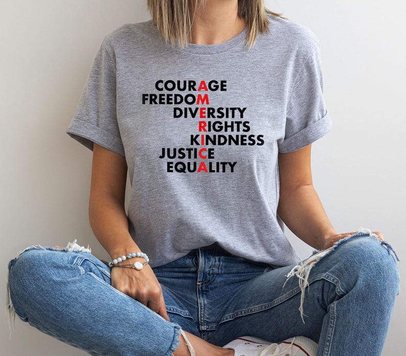 May include: A light gray t-shirt with the words "COURAGE, FREEDOM, DIVERSITY, RIGHTS, KINDNESS, JUSTICE, EQUALITY" printed in black and red. The shirt is worn with blue jeans.