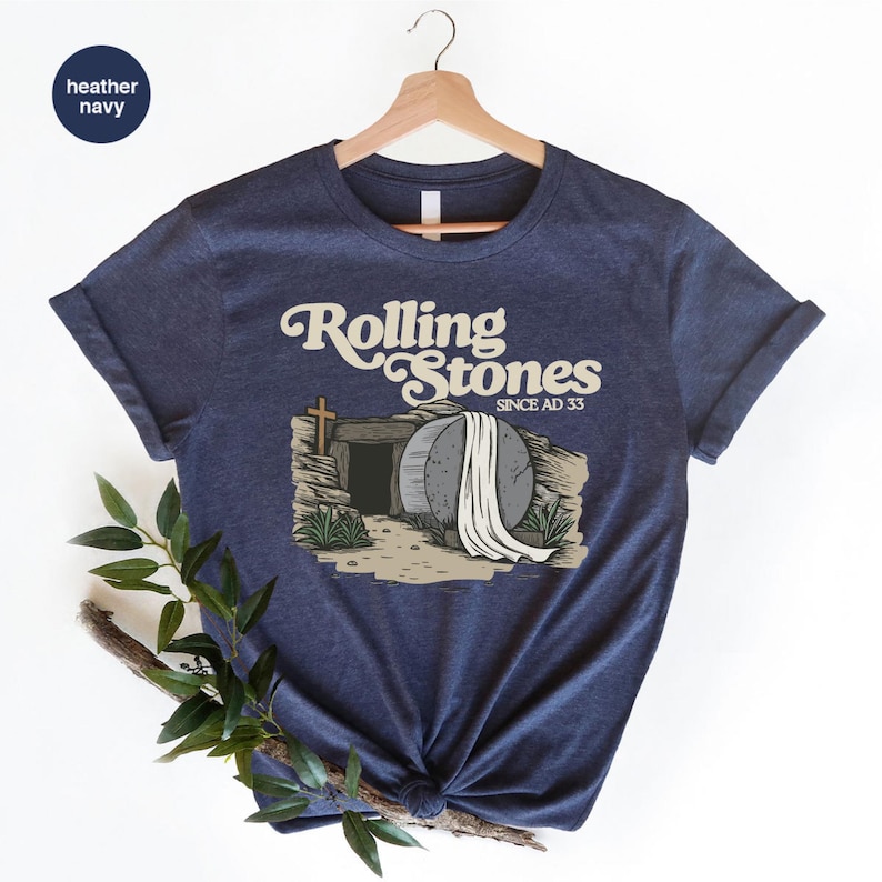 May include: A heather navy blue t-shirt with a graphic of a stone tomb with a white sheet draped over it. The text "Rolling Stones Since AD 33" is above the tomb.
