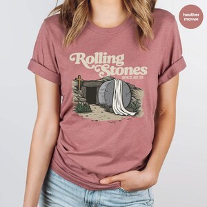 May include: Heather mauve t-shirt with a graphic of a stone tomb with a white sheet draped over it. The text "Rolling Stones Since AD 33" is above the tomb.