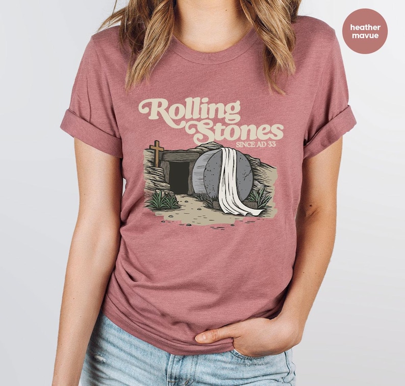 May include: Heather mauve t-shirt with a graphic of a stone tomb with a white sheet draped over it. The text "Rolling Stones Since AD 33" is above the tomb.