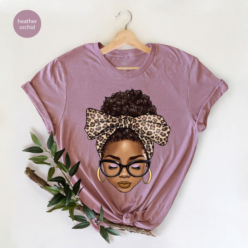 May include: A heather orchid t-shirt with a graphic of a woman with a brown bun, wearing a leopard print headband and glasses.
