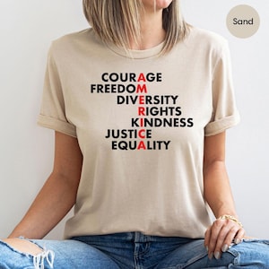 May include: Sand-colored t-shirt with the word "AMERICA" spelled vertically using words like "Courage," "Freedom," and "Equality." The letters "A," "R," and "A" are in red. The shirt is made of a soft material.
