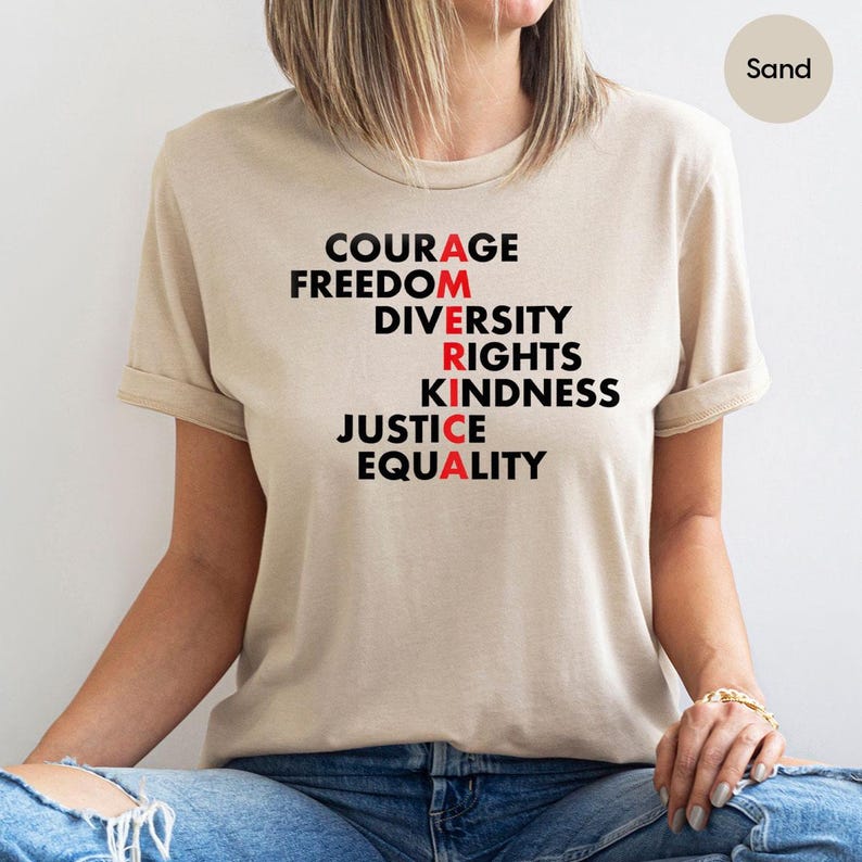 May include: Sand-colored t-shirt with the word "AMERICA" spelled vertically using words like "Courage," "Freedom," and "Equality." The letters "A," "R," and "A" are in red. The shirt is made of a soft material.