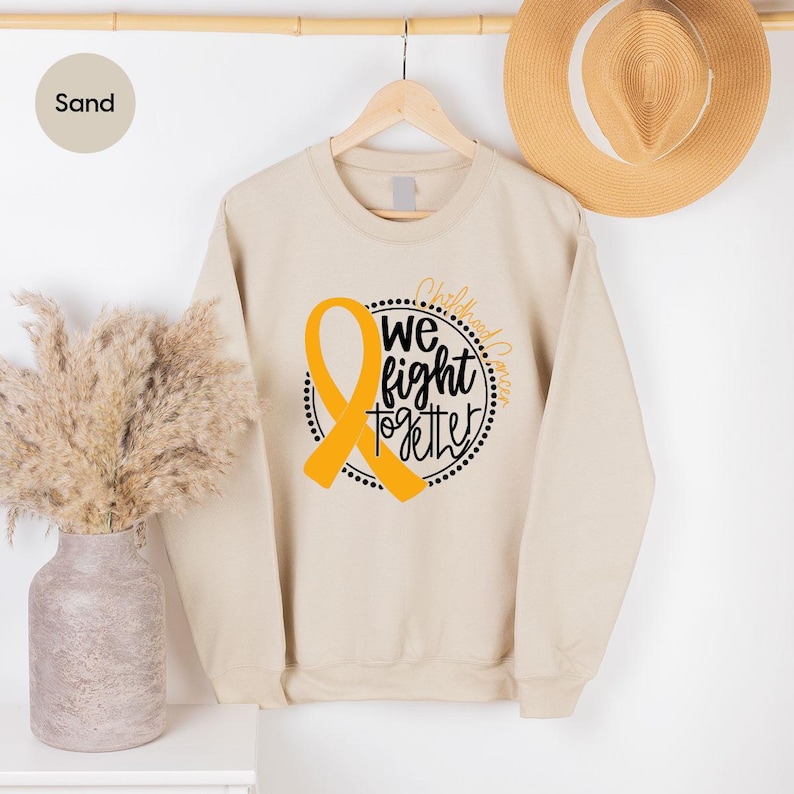 May include: Sand colored sweatshirt with a gold ribbon and the text "We fight together" in a black circle with the words "Childhood Cancer" above the circle.