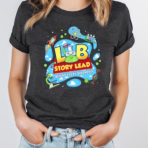 May include: Dark grey t-shirt with a vibrant graphic. The design features the text "LB STORY LEAD BEHIND EVERY DIAGNOSIS" alongside illustrations of scientific elements, including a rocket, microscope, and DNA strand.