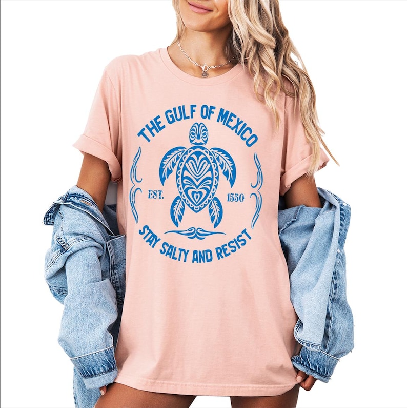 Puede incluir: Una camiseta rosa con un gr&aacute;fico azul de una tortuga marina con el texto "The Gulf of Mexico Est. 1550 Stay Salty and Resist".