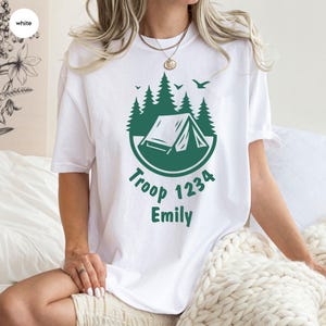 Może przedstawiać: Biała koszulka z zielonym motywem kempingowym. Projekt zawiera namiot, drzewa i ptaki, z tekstem "Troop 1234 Emily" w okrągłym układzie. Koszulka jest w stylu casual z kr&oacute;tkim rękawem.