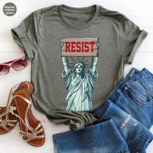 May include: Heather military green t-shirt with a graphic of the Statue of Liberty holding a sign with the word "RESIST" in red. The image is accompanied by blue denim jeans, brown sandals, and pink sunglasses.
