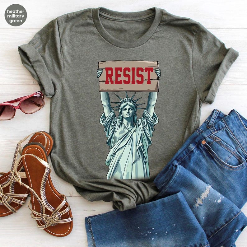 May include: Heather military green t-shirt with a graphic of the Statue of Liberty holding a sign with the word "RESIST" in red. The image is accompanied by blue denim jeans, brown sandals, and pink sunglasses.