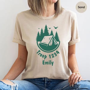 Może przedstawiać: Beżowa koszulka z zieloną grafiką o tematyce kempingowej. Projekt zawiera namiot, drzewa i tekst "Troop 1234 Emily". Koszulka ma kr&oacute;tkie rękawy i okrągły dekolt.