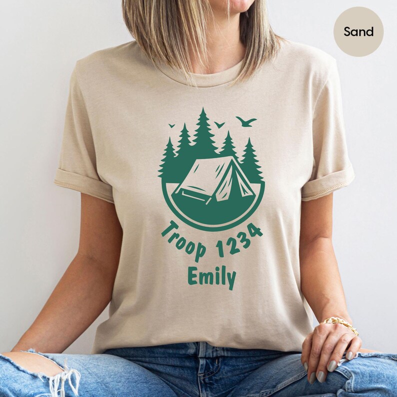 Może przedstawiać: Beżowa koszulka z zieloną grafiką o tematyce kempingowej. Projekt zawiera namiot, drzewa i tekst "Troop 1234 Emily". Koszulka ma kr&oacute;tkie rękawy i okrągły dekolt.