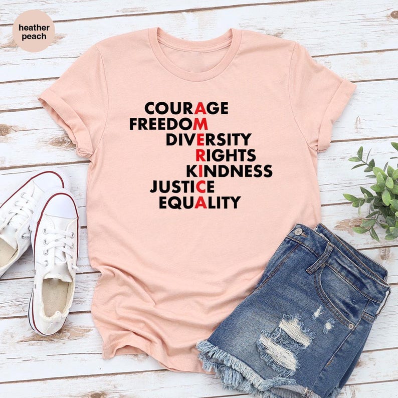 May include: A light peach-colored t-shirt with the words "COURAGE, FREEDOM, DIVERSITY, RIGHTS, KINDNESS, JUSTICE, EQUALITY" printed in black and red. The shirt is paired with denim shorts and white canvas sneakers.