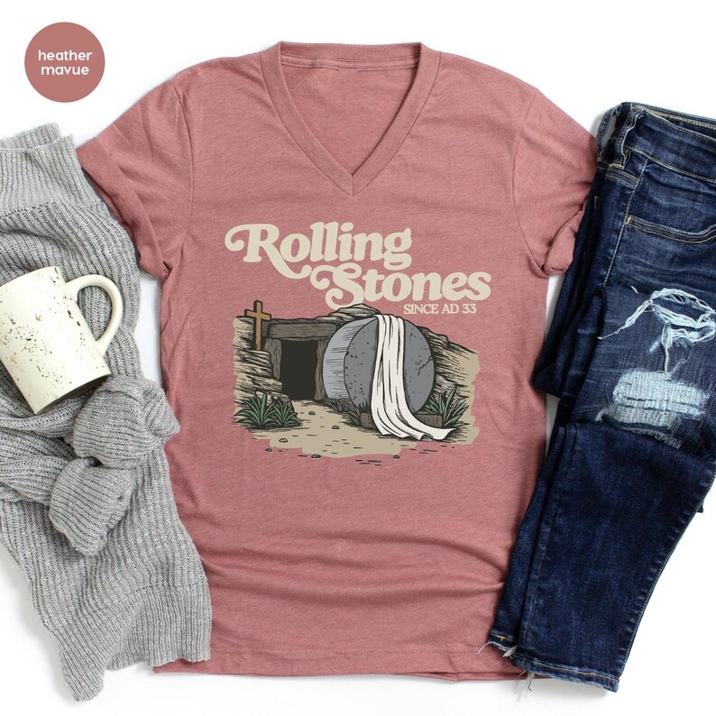 May include: A heather mauve v-neck t-shirt with a graphic of a stone tomb with a white sheet and the text "Rolling Stones Since AD 33".