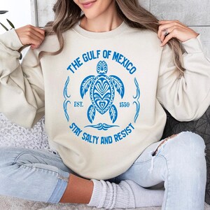 Puede incluir: Una sudadera de color crema con un gr&aacute;fico azul de una tortuga marina y el texto "The Gulf of Mexico Est. 1550 Stay Salty and Resist".