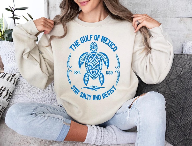Puede incluir: Una sudadera de color crema con un gr&aacute;fico azul de una tortuga marina y el texto "The Gulf of Mexico Est. 1550 Stay Salty and Resist".