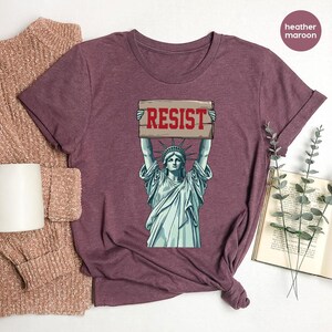May include: Heather maroon t-shirt with a graphic of the Statue of Liberty holding a sign with the word "RESIST" in red. The shirt has short sleeves and a crew neck. The fabric appears soft and comfortable.