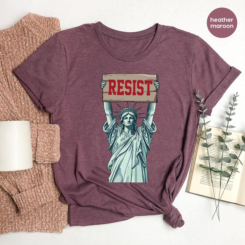 May include: Heather maroon t-shirt with a graphic of the Statue of Liberty holding a sign with the word "RESIST" in red. The shirt has short sleeves and a crew neck. The fabric appears soft and comfortable.