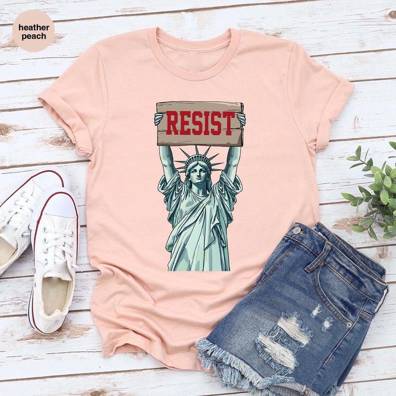 May include: A pale peach t-shirt with a graphic of the Statue of Liberty holding a sign with the word "RESIST" in red. The shirt is styled with denim shorts and white canvas trainers.