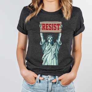 May include: Dark grey t-shirt with a graphic of the Statue of Liberty holding a sign with the word "RESIST" in red. The Statue of Liberty is in light blue. The t-shirt has rolled-up sleeves.