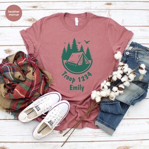 Może przedstawiać: R&oacute;żowa koszulka z zieloną grafiką kempingową i napisem "Troop 1234 Emily". Koszulka jest połączona z białymi trampkami, szalikiem w kratę i niebieskimi dżinsami. Koszulka ma okrągły dekolt i kr&oacute;tkie rękawy.