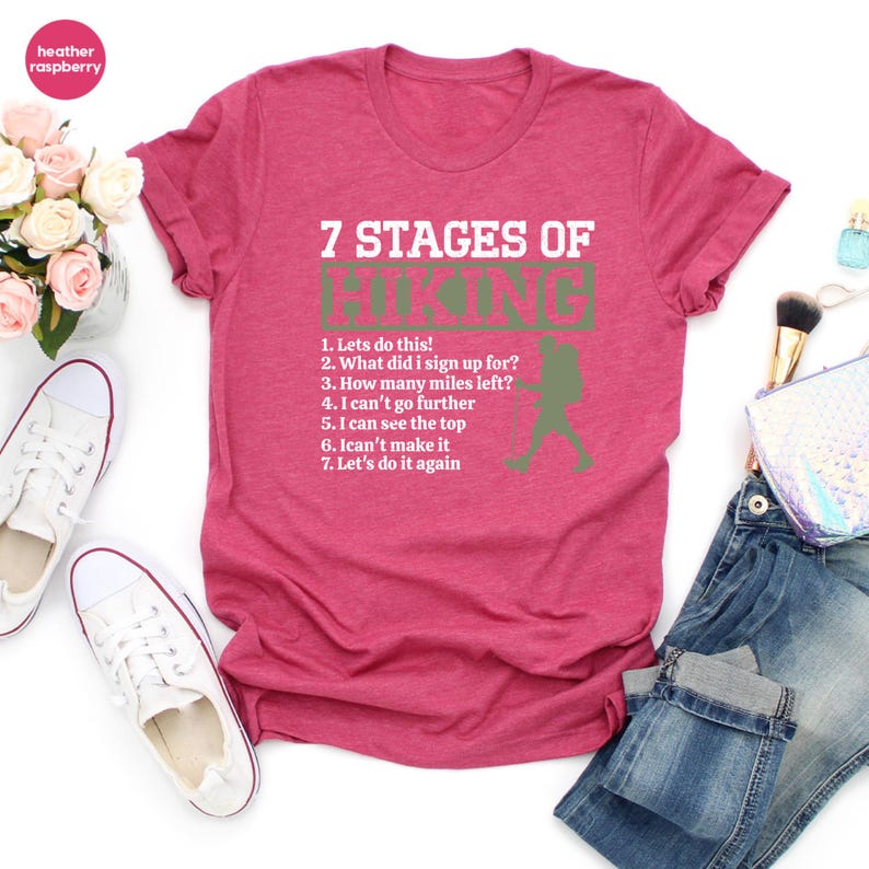 May include: A heather raspberry short-sleeve t-shirt featuring white text that reads 7 STAGES OF HIKING, followed by a numbered list of seven hiking-related phrases and a green silhouette of a hiker with a backpack and trekking poles.
